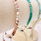 Three crystal beaded More Love Meridian bracelets displayed on a cream fabric jewelry stand. One bracelet is made of light pink round beads with metallic accents called Rhodonite Crystal, one is made of turquoise-blue rectangular beads called Navajo Turquoise, and one is a delicate Turquoise crystal bracelet with small beads and dangling charms. Metaphysical jewelry to spread love world wide.