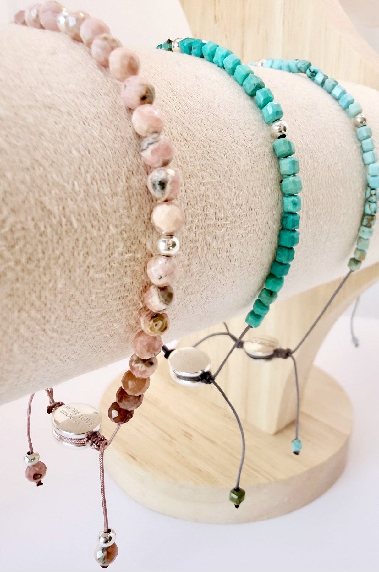 Three crystal beaded More Love Meridian bracelets displayed on a cream fabric jewelry stand. One bracelet is made of light pink round beads with metallic accents called Rhodonite Crystal, one is made of turquoise-blue rectangular beads called Navajo Turquoise, and one is a delicate Turquoise crystal bracelet with small beads and dangling charms. Metaphysical jewelry to spread love world wide.